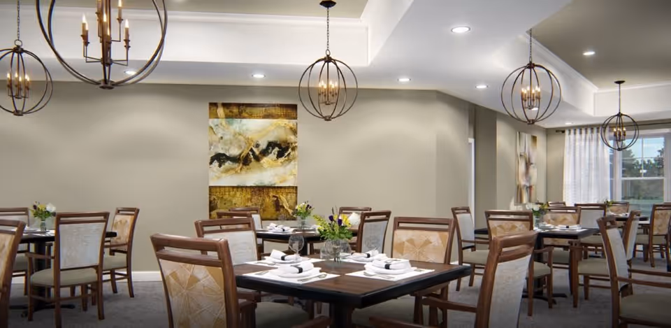 Well-lit communal dining room with multiple set tables and wooden chairs, modern spherical chandeliers, wall artwork and windows.
