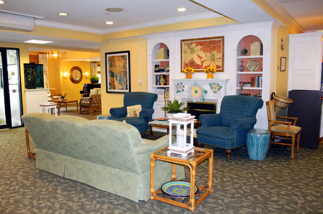 A cozy assisted living facility common area with a green patterned sofa, two blue armchairs, a wooden side table with a white lantern, and a fireplace decorated with colorful flower crafts and sunflowers. The walls are painted yellow and white with built-in shelves holding books and decorative items. There is a carpeted floor and additional seating visible in the background near a glass door entrance.
