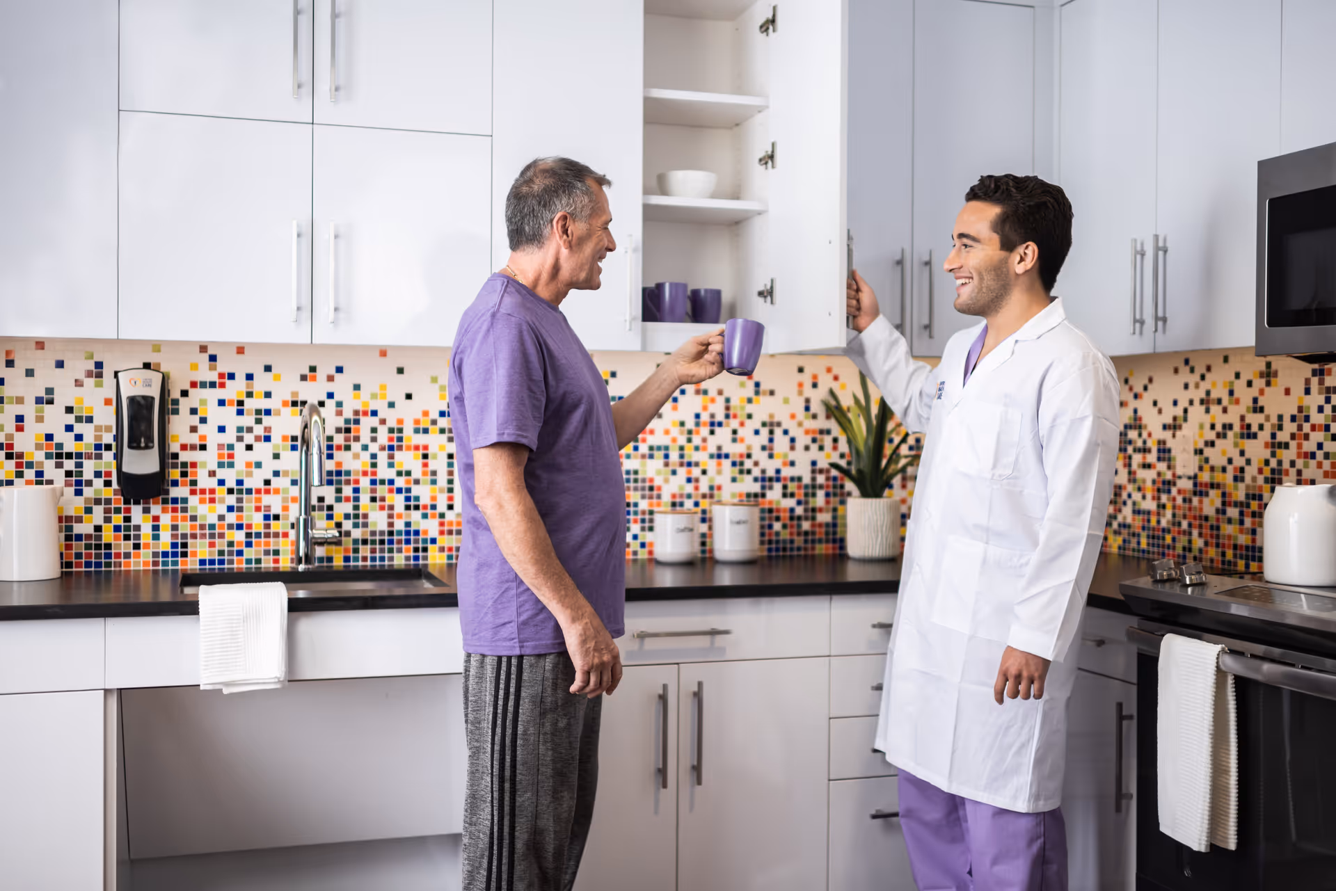 A man in a purple shirt and gray pants holds a purple mug while talking to a healthcare professional in a white coat and purple scrubs in a modern kitchen with white cabinets and a colorful mosaic tile backsplash.