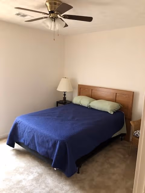 A simple bedroom with a double bed covered in a blue bedspread, a wooden headboard, bedside table with lamp, and a ceiling fan.