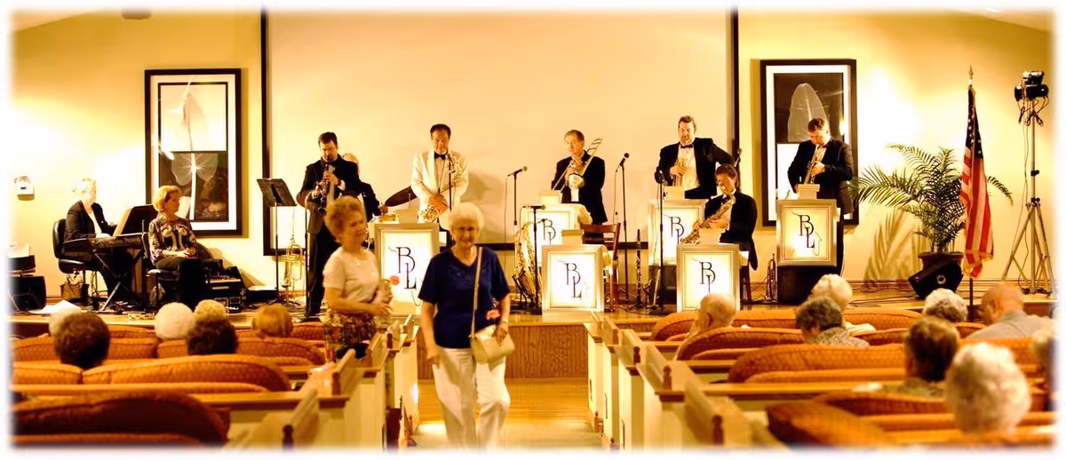 A live band performing on a stage in front of an audience seated in rows of chairs. Two elderly women are walking down the aisle. The band consists of musicians playing various instruments including saxophones, trumpet, trombone, and keyboard. The setting appears to be an indoor community or event room with warm lighting and framed artwork on the walls.