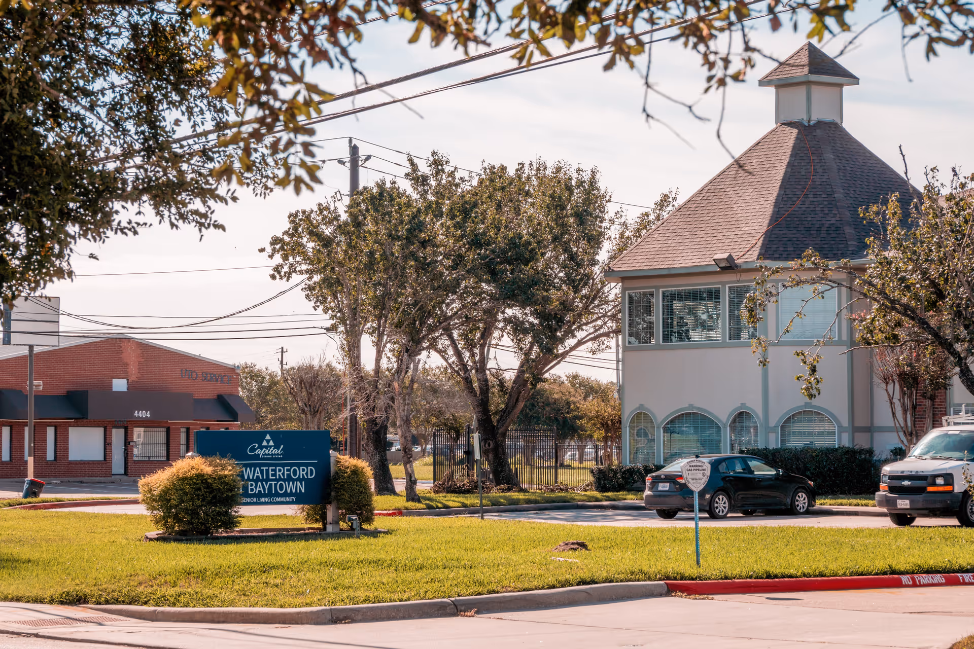 Exterior view of The Waterford at Baytown senior living community showing a building with a unique roof design, a green lawn, trees, parked cars, and a sign with the facility's name.