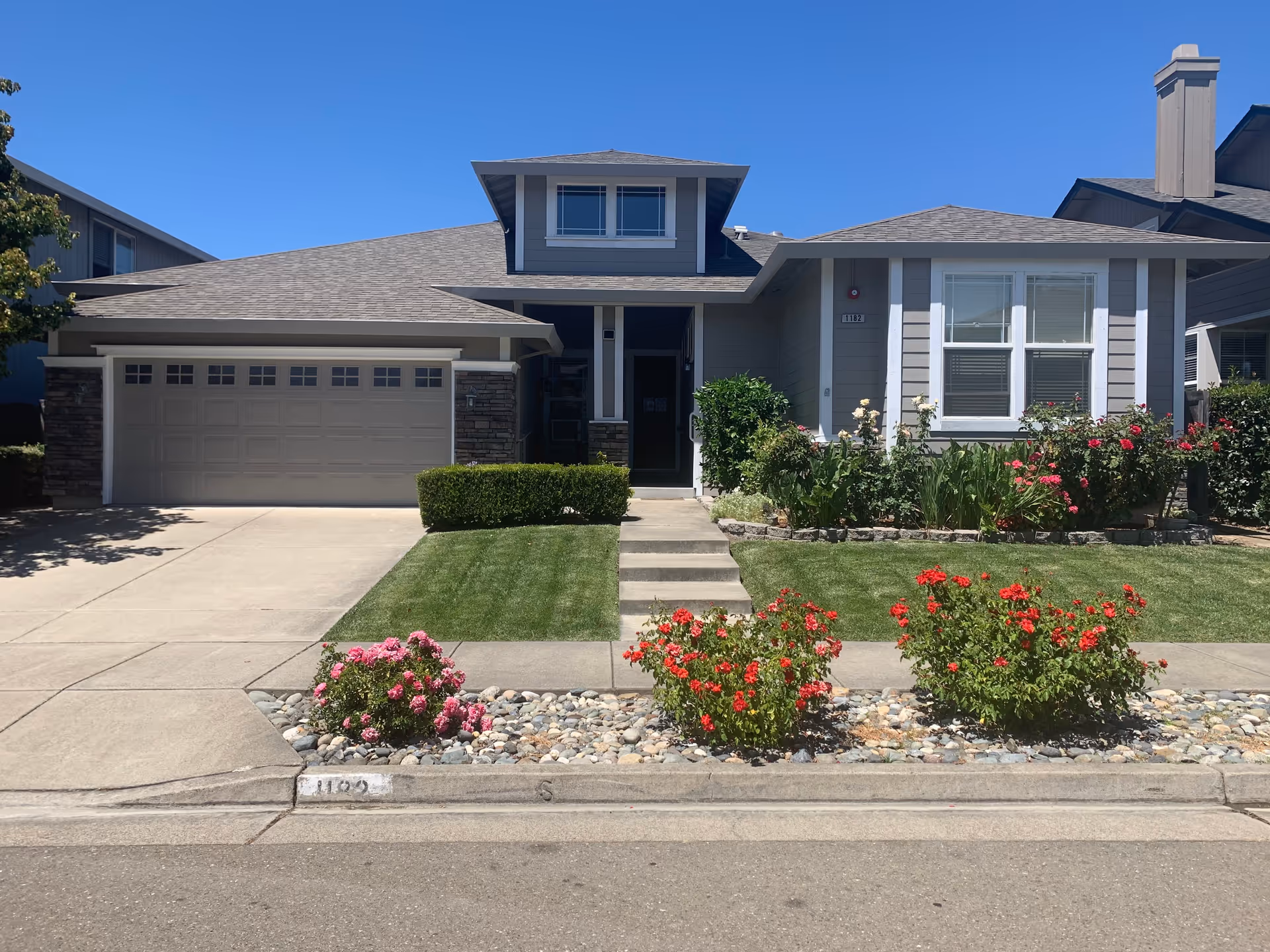 Front exterior view of a single-story house with a two-car garage, a small front porch, well-maintained lawn, and flower beds with red and pink flowers under a clear blue sky.