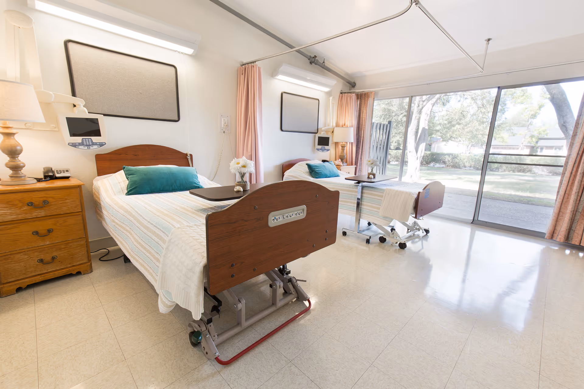 A bright and clean room with two hospital-style beds, each with a green pillow and a small table on wheels. There are bedside tables with lamps and medical equipment mounted on the wall. Large sliding glass doors open to a view of trees and greenery outside, allowing natural light to fill the room.