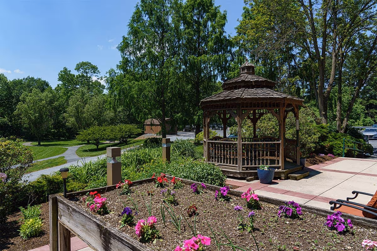 Wooden gazebo next to a raised flower bed and paved walkway in a landscaped outdoor garden with trees.