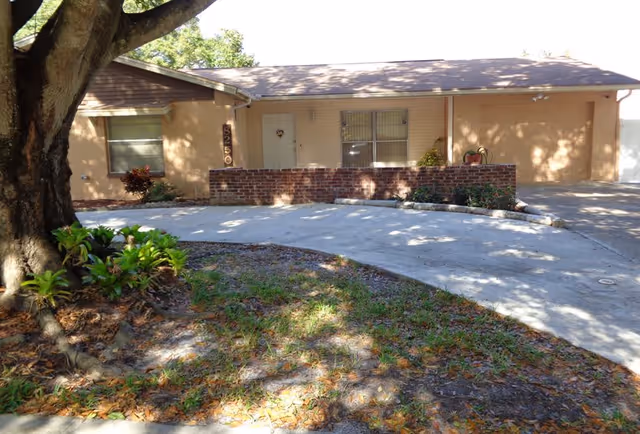 Front exterior view of a single-story residential building with a large tree and some plants in the foreground. The building has a beige exterior, a brick wall near the entrance, a driveway, and a garage on the right side.