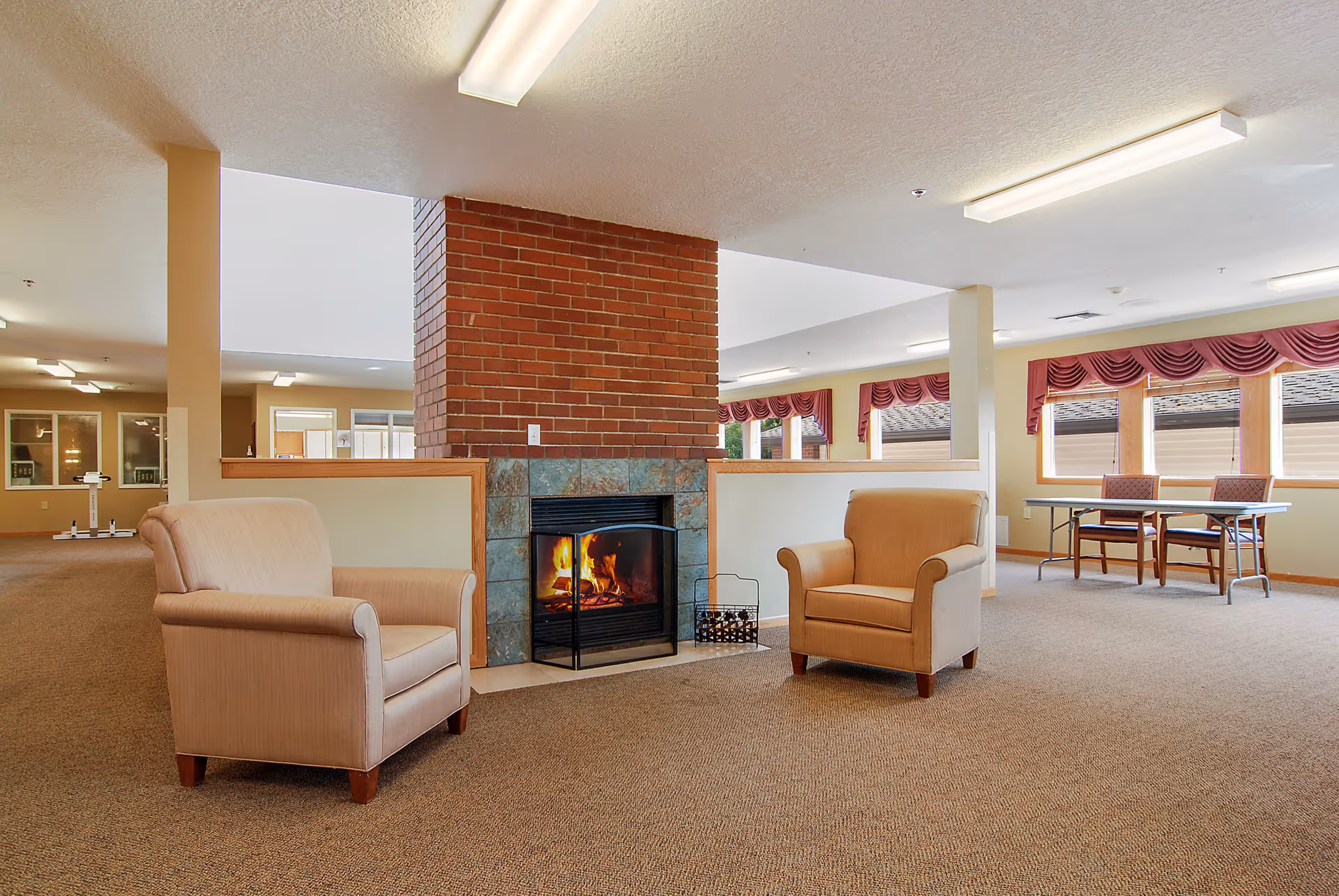 A cozy common area with two beige armchairs facing a brick fireplace with a fire burning. The room has carpeted floors, large windows with red valances, and fluorescent ceiling lights. A table with two chairs is visible near the windows.