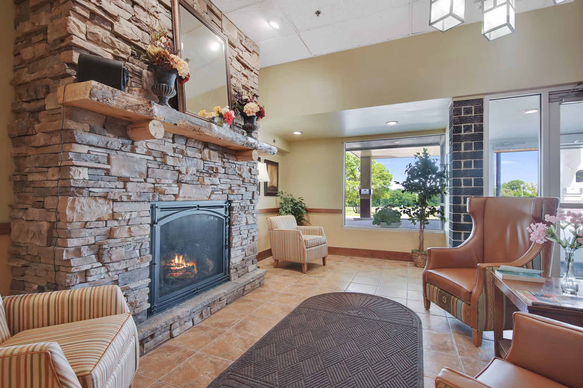 Cozy lounge area with a stone fireplace, upholstered chairs, and a large window overlooking greenery.