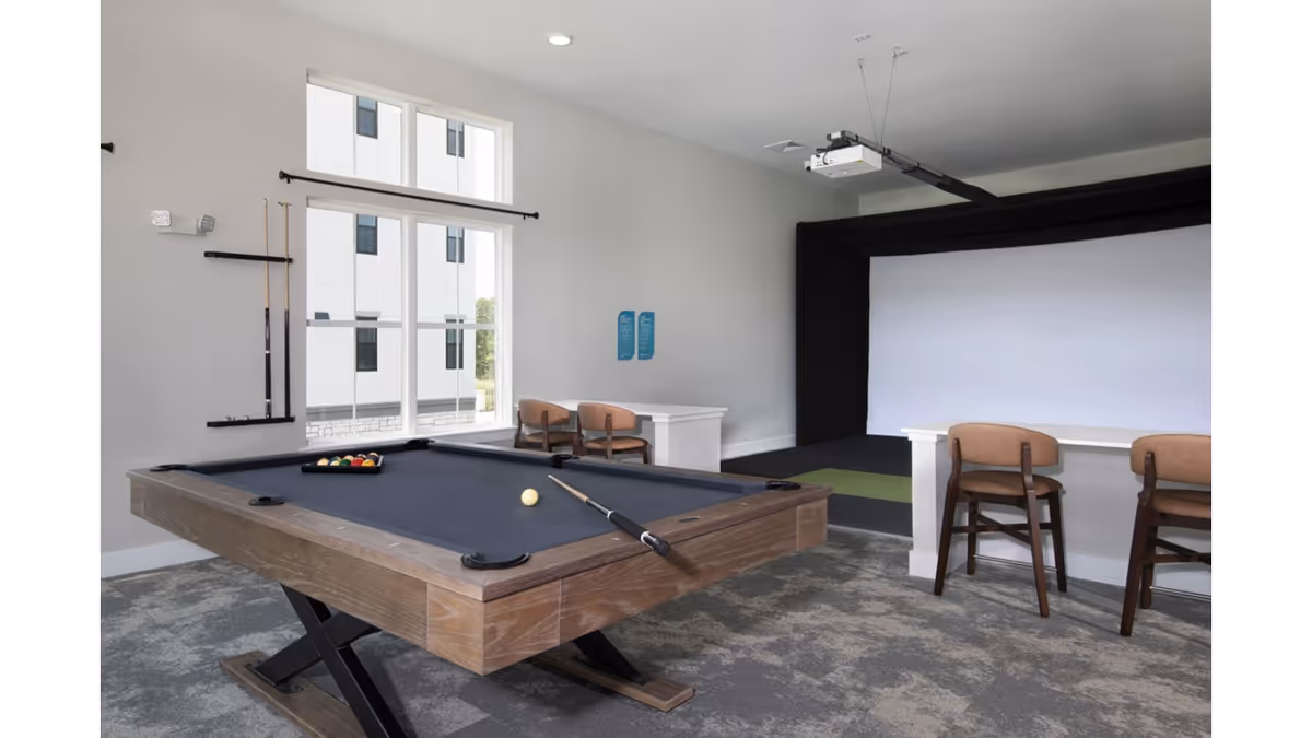 A recreational room with a pool table in the foreground, a cue stick resting on it, and a set of pool balls arranged for a game. Behind the pool table, there are two chairs and a counter or bar area. On the far wall, there is a large white projection screen with a projector mounted on the ceiling. The room has large windows letting in natural light and a carpeted floor.