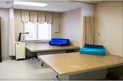 A therapy or treatment room with two padded treatment tables, each with a rolled-up exercise mat on top. The room has a window with blinds and a beige curtain divider on one side. The walls and floor are light-colored, creating a clean and clinical environment.