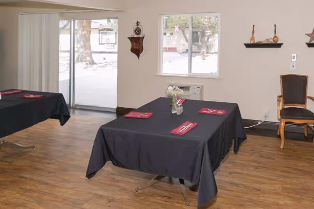Interior dining area with two tables covered in black tablecloths, each set with red napkins and silverware. The room has wooden flooring, a window, a sliding glass door, and a single wooden chair against the wall. Decorative items are mounted on the walls.