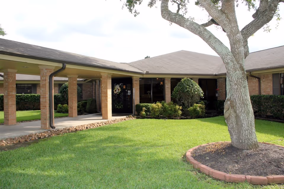 Exterior view of Baywind Village Skilled Nursing and Rehabilitation showing a single-story brick building with a covered walkway supported by brick columns. There is a well-maintained lawn with a tree and landscaped bushes near the entrance door decorated with a wreath.