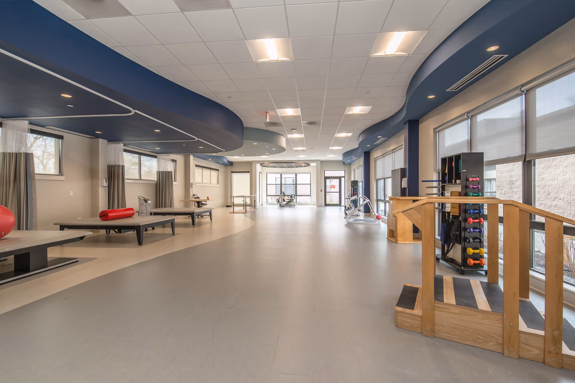 A spacious rehabilitation or physical therapy room with exercise equipment including therapy tables, a set of stairs with handrails, and a rack of colorful dumbbells. The room has large windows with blinds, a curved blue ceiling feature, and a light gray floor.