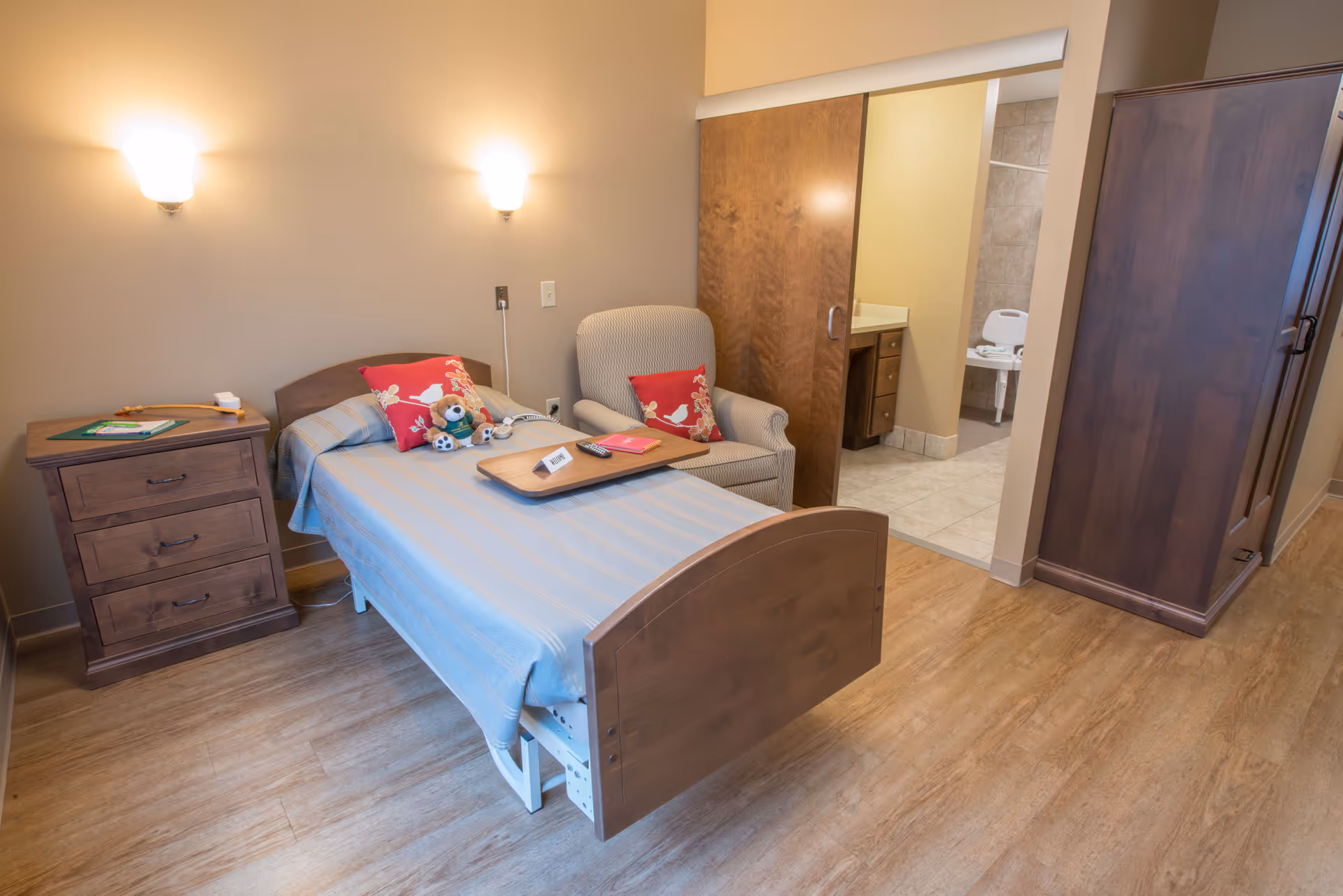 A cozy bedroom in a senior living facility featuring a single bed with a blue striped blanket and decorative pillows, a wooden nightstand with drawers, a comfortable armchair with a red pillow, and a sliding door leading to a bathroom with tiled floors and a shower chair.