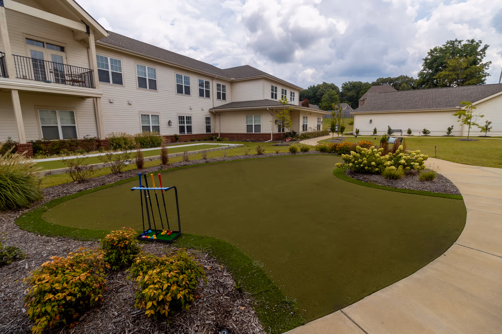 Courtyard of a senior living facility featuring a small artificial putting green with a croquet set, landscaped beds, and surrounding two-story buildings.