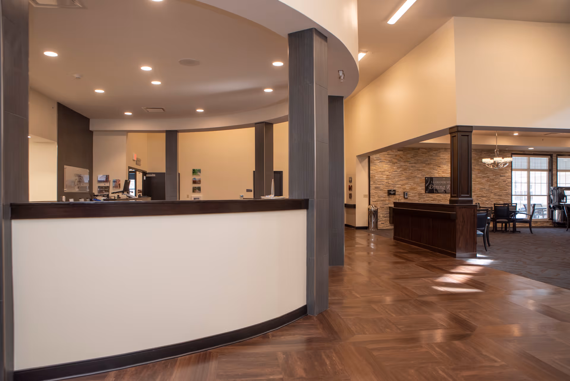 Interior view of a senior living facility reception area with a curved white and dark wood front desk. The space has warm lighting, wood-patterned flooring, and a dining room area visible in the background with tables and chairs near large windows.