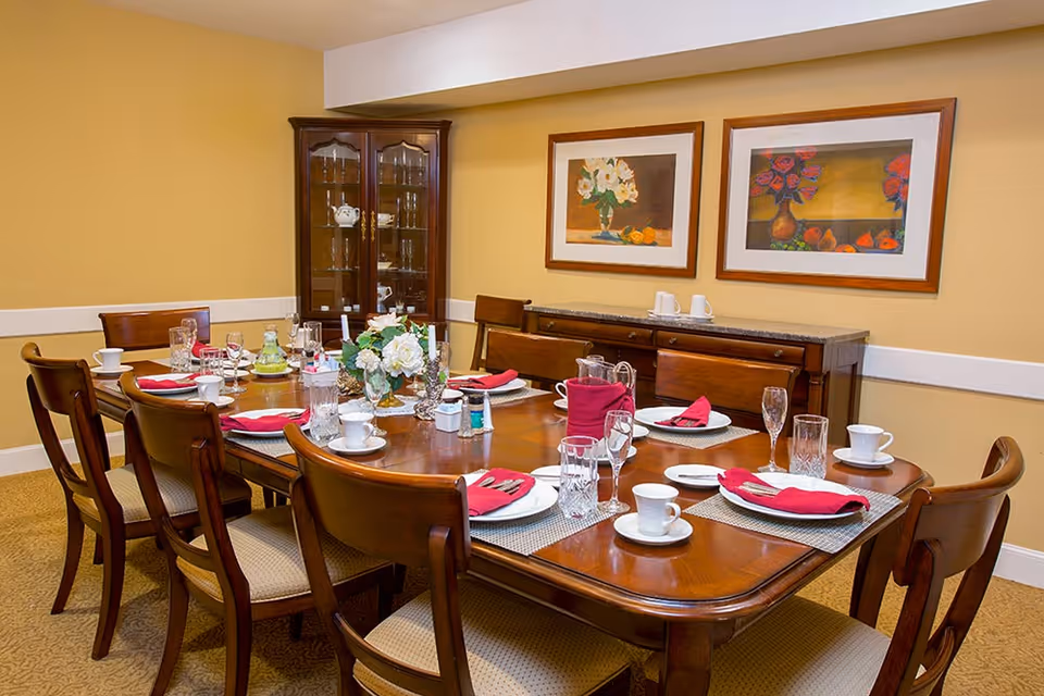 A formal dining room with a wooden dining table set for eight people. The table is arranged with white plates, cups, glasses, silverware, and red cloth napkins. A floral centerpiece and salt and pepper shakers are in the middle of the table. Behind the table is a wooden sideboard with two framed floral paintings hanging above it, and a glass-fronted china cabinet in the corner.