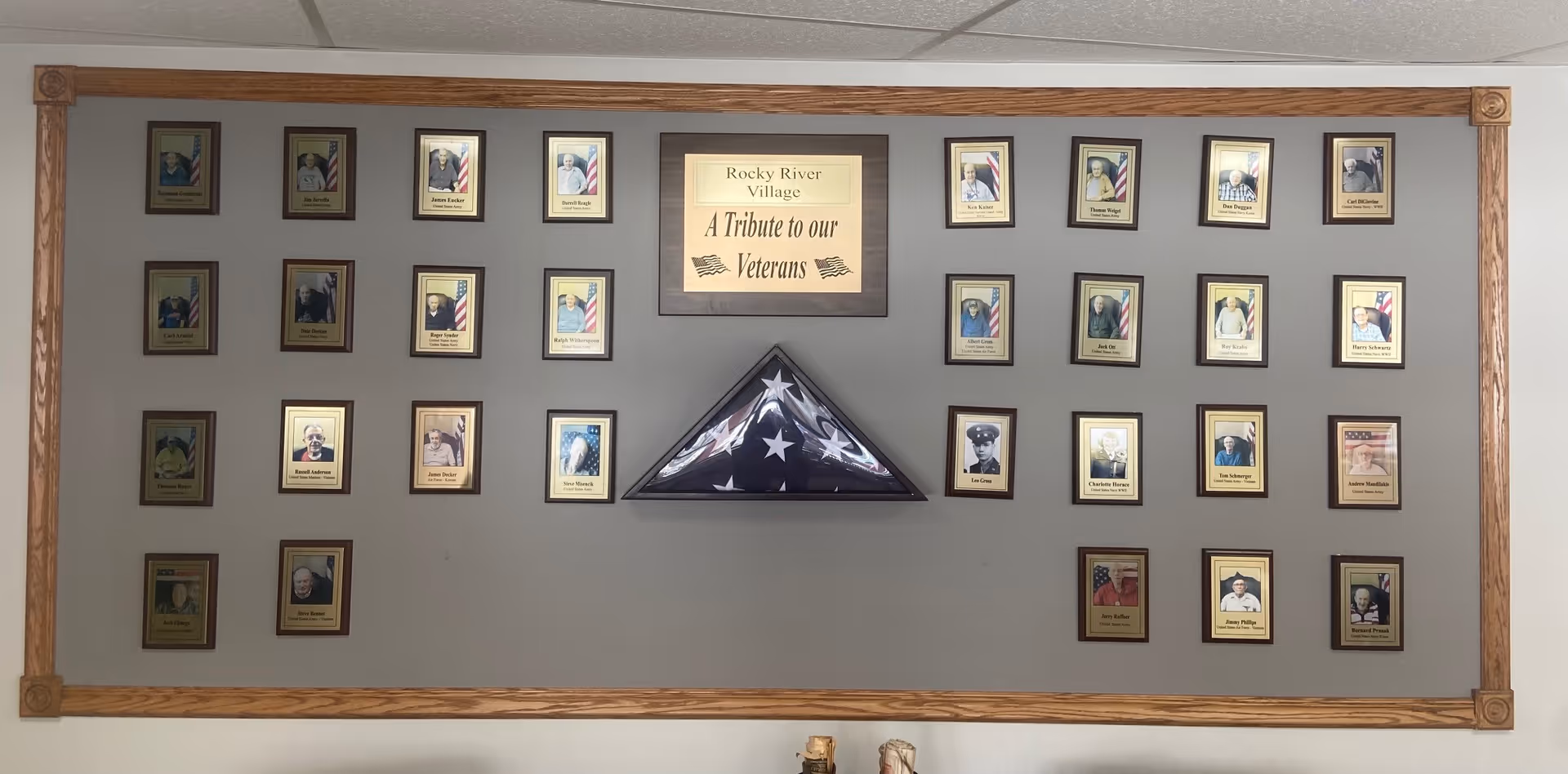 A tribute wall at Rocky River Village featuring framed photos of veterans arranged around a central plaque that reads 'Rocky River Village A Tribute to our Veterans' and a folded American flag in a triangular display case, all mounted on a gray wall with wooden trim.
