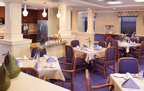 A dining room in an assisted living facility with tables covered in white tablecloths, each set with purple napkins, plates, cups, and silverware. The room features purple carpet and chairs with purple upholstery. There is a kitchen area with stainless steel appliances and pendant lights in the background. A popcorn machine is visible near the windows.