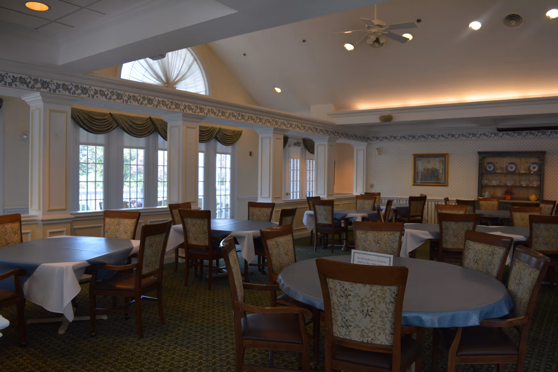 A dining room with multiple tables covered with tablecloths and surrounded by wooden chairs with patterned upholstery. The room has large windows with green valances, a high ceiling with recessed lighting, and a decorative cabinet with plates on display against the far wall.
