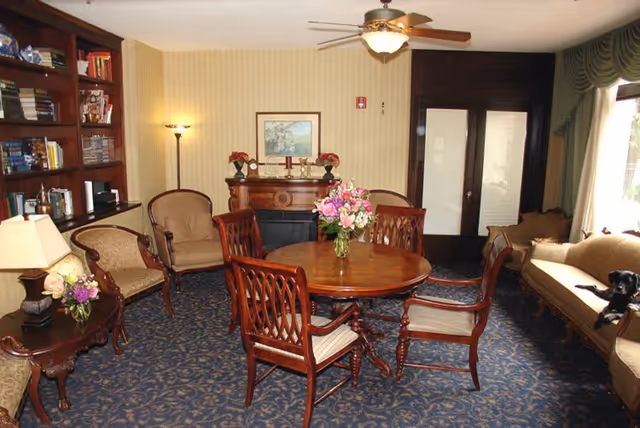 A cozy living room with a round wooden table surrounded by four wooden chairs with cushions. There is a floral centerpiece on the table. The room features a blue patterned carpet, a beige sofa with a black dog sitting on it, two armchairs, a wooden side table with a lamp and flowers, a tall floor lamp, a wooden bookshelf filled with books, and a wooden fireplace mantel with decorative items and a framed picture above it. The room has beige striped wallpaper and a ceiling fan with a light fixture. French doors with frosted glass panels are visible in the background.