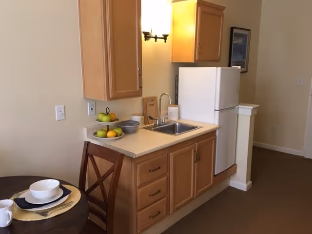 Small kitchenette with light wood cabinets, a sink and refrigerator beside a small dining table set with bowls and a fruit plate.
