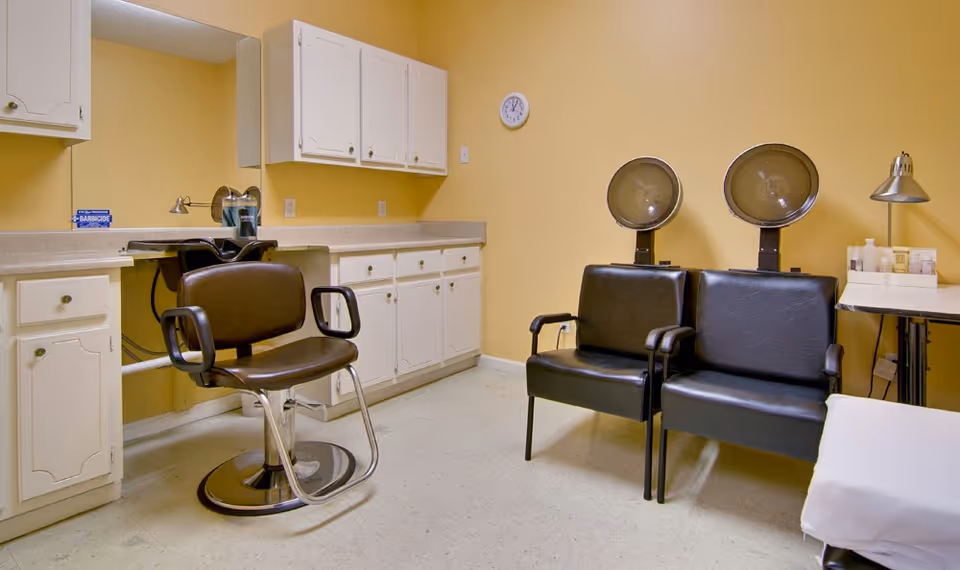 Interior of a senior living facility's hair salon area with a brown salon chair in front of a large mirror, white cabinets along the wall, two black chairs with hair dryers, a small table with hair products, and a wall clock.