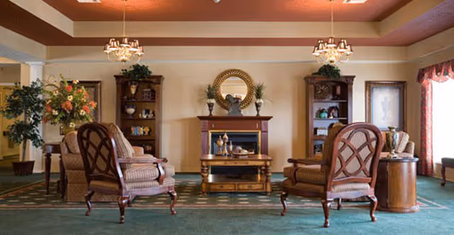 A cozy living room area with four upholstered armchairs arranged around a wooden coffee table in front of a fireplace. The fireplace has a decorative round mirror and two vases on the mantel. Two wooden bookshelves flank the fireplace, each with decorative items and plants on top. The room features green carpet, beige walls, and two chandeliers hanging from the ceiling. A large window with curtains is visible on the right side.