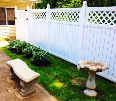 Outdoor area at a senior facility featuring a curved stone bench, birdbath, white fence, and greenery.
