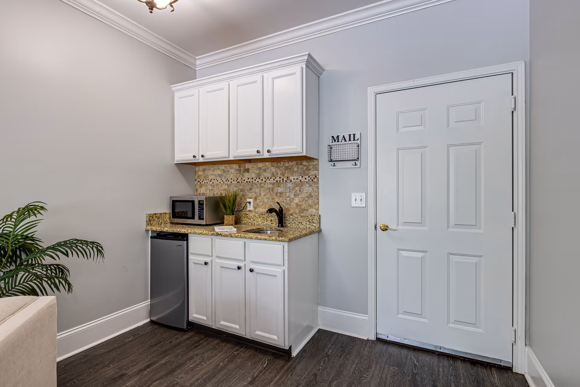 Small kitchenette area with white cabinets, granite countertop, a small stainless steel refrigerator, a microwave, a small sink with a black faucet, and a decorative plant. There is a door to the right and a mail organizer mounted on the wall above the light switch. The floor is dark wood, and a green plant is partially visible on the left side.