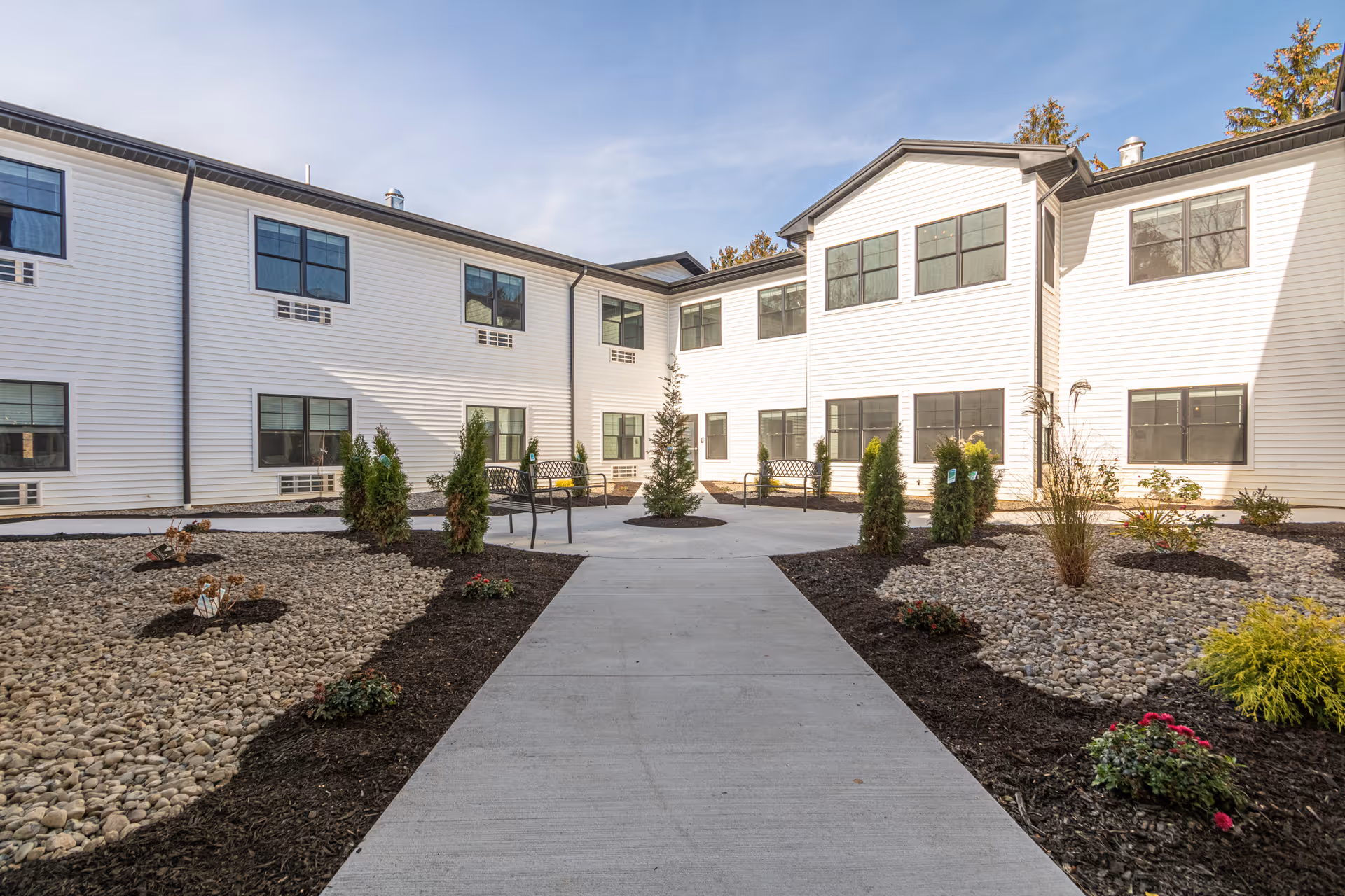 Outdoor courtyard area of a senior living facility with a concrete pathway leading to a central circular area with benches and small trees. The building surrounding the courtyard is two stories tall with white siding and multiple windows. The landscaping includes mulch, rocks, and small plants.