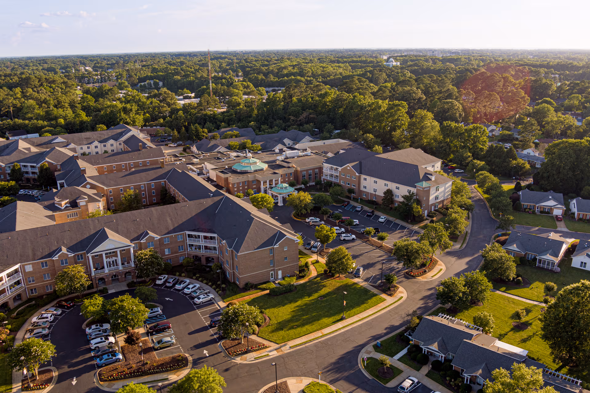 Aerial view of The Chesapeake senior living facility showing multiple connected buildings with parking lots, surrounded by trees and residential houses under a clear sky.