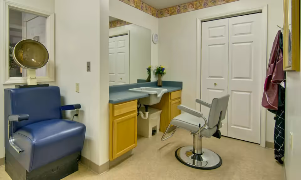 Interior view of a salon area in a senior living facility featuring a blue salon chair with a hair dryer hood, a gray salon chair in front of a sink with a large mirror above, wooden cabinets below the sink, and a closet with white double doors. A floral wallpaper border runs along the top of the walls.