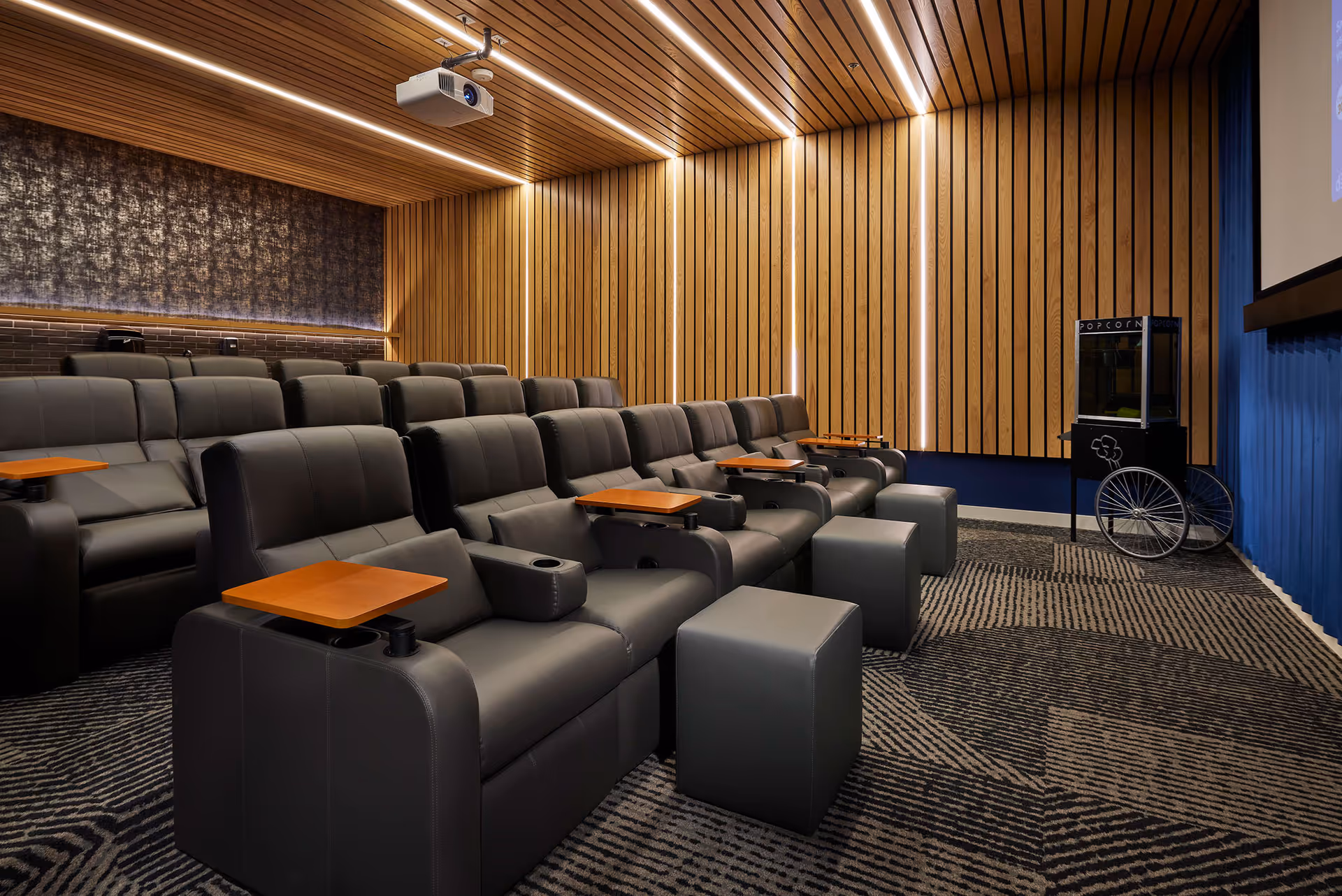 A modern theater room with multiple rows of black leather reclining seats, each equipped with a small wooden tray table. The room features wood paneling on the walls and ceiling, a patterned carpet, a popcorn machine on the right side, and a projector mounted on the ceiling.