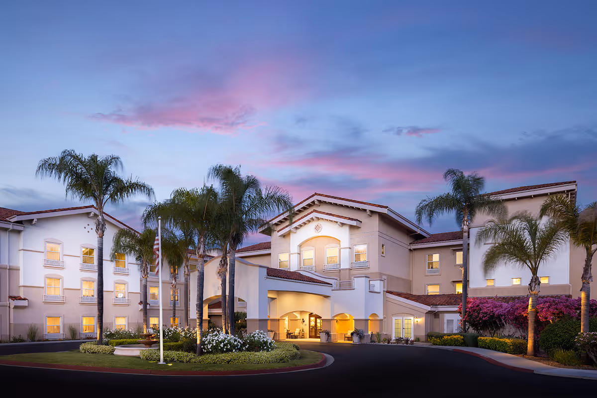 Exterior view of Belmont Village Senior Living Sabre Springs building at dusk with palm trees, landscaped garden, and a circular driveway under a colorful sky.