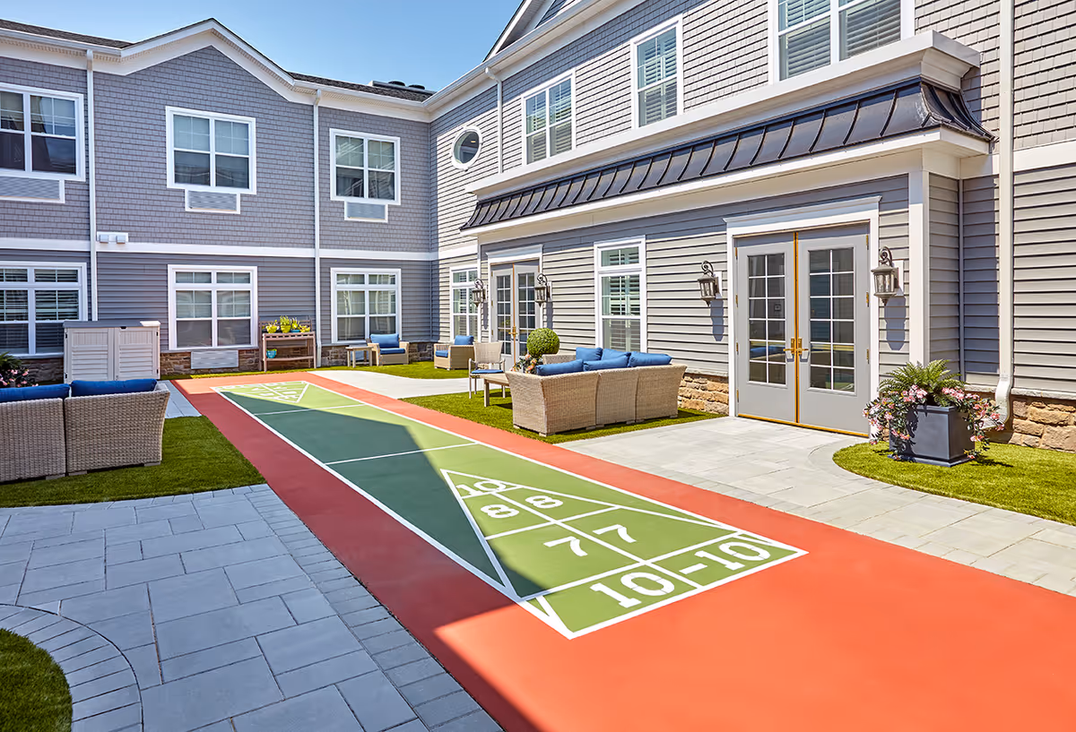 Outdoor courtyard area of The Bristal Assisted Living at Mount Sinai featuring a shuffleboard court, patio seating with wicker furniture and blue cushions, potted plants, and a two-story building with multiple windows and French doors.