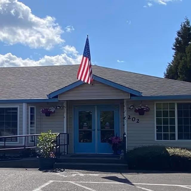 Front exterior view of a single-story building with a gray roof and light-colored siding. The entrance has double blue doors with glass panels, flanked by two windows. An American flag is mounted above the entrance. There are hanging flower baskets on either side of the door and potted plants near the entrance. The building number 4202 is visible on the right side of the door. The sky is partly cloudy with blue patches.