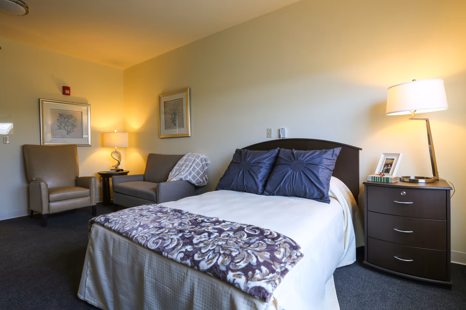 A cozy senior living bedroom with a neatly made bed featuring two dark blue pillows and a patterned blanket at the foot. To the right of the bed is a dark wooden nightstand with a modern lamp, a framed photo, and a book. On the left side of the room, there is a comfortable armchair and a small sofa with a throw blanket. The walls are decorated with two framed botanical prints, and the room is softly lit by two lamps.