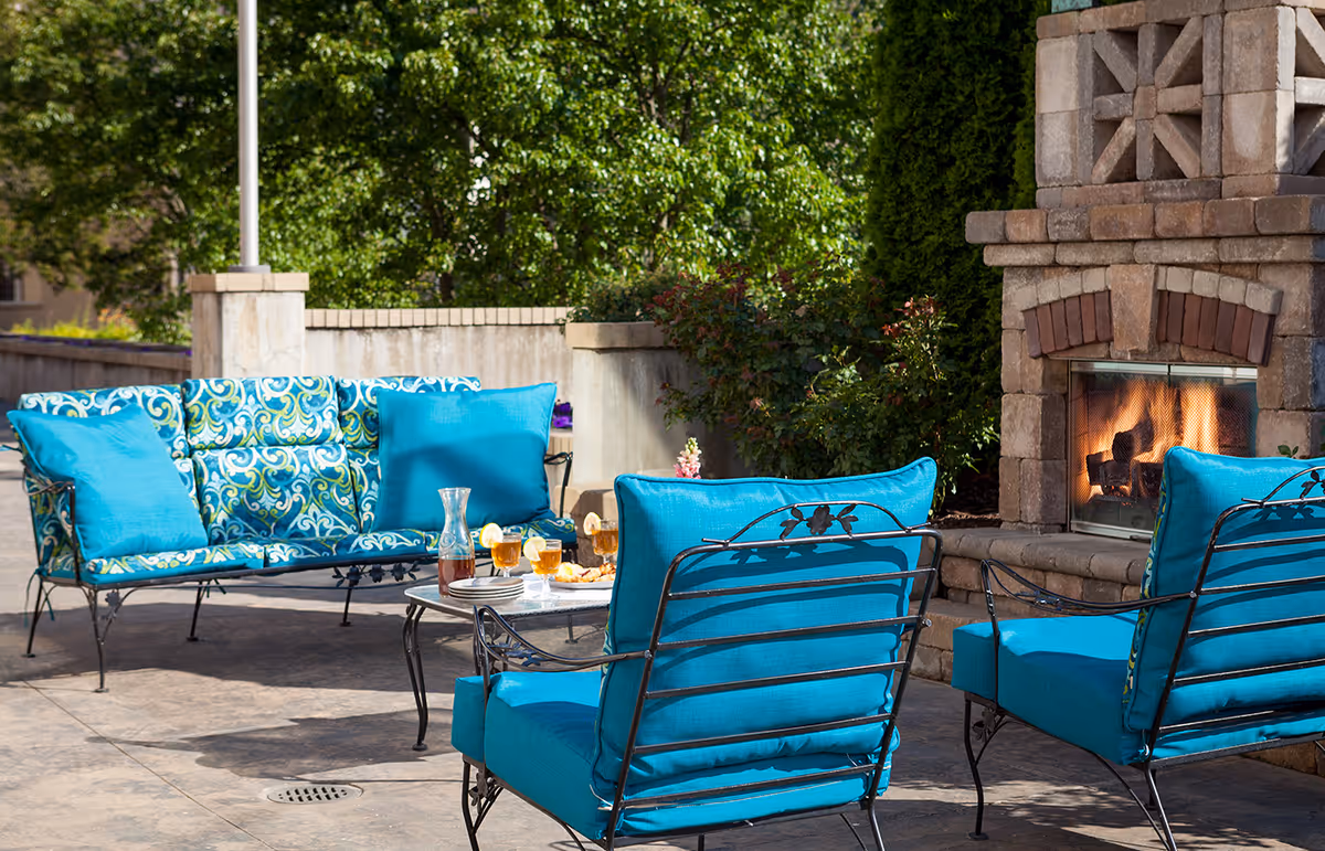 Outdoor patio area with blue cushioned metal chairs and a matching loveseat arranged around a small table with drinks and snacks. A stone fireplace with a fire burning is visible on the right side, and green trees and bushes are in the background.