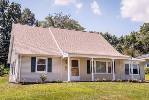 Single-story house with beige siding, a steeply pitched roof, black shutters, and a front porch with white columns. The house is surrounded by a well-maintained lawn and some small shrubs, with trees and a partly cloudy sky in the background.