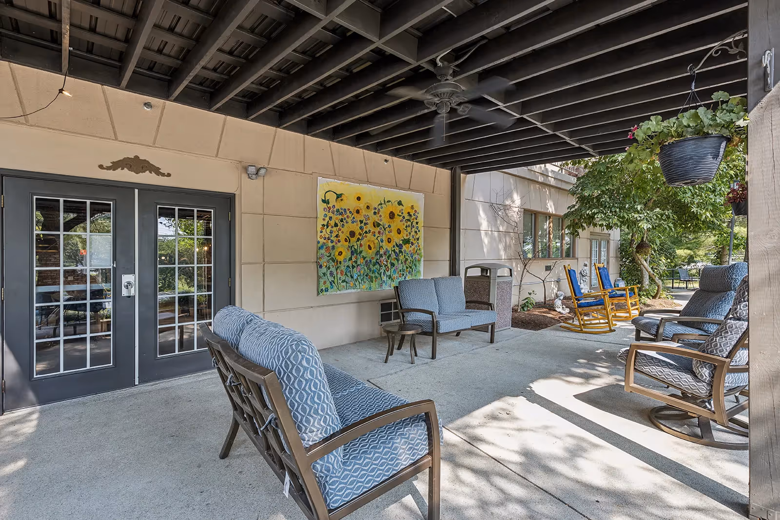 Covered outdoor patio area with cushioned seating including chairs and a loveseat, a ceiling fan, hanging plants, and a colorful sunflower painting on the wall. There are also two wooden rocking chairs and a trash bin near the wall.