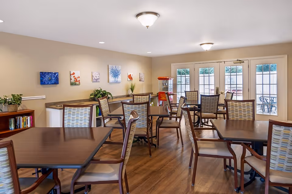 A bright dining room with several wooden tables and cushioned chairs arranged neatly. The room has light beige walls decorated with colorful floral paintings and some potted plants on a counter along one wall. There is a popcorn machine near glass double doors that lead outside to a patio area. The floor is wooden, and the ceiling has multiple light fixtures.
