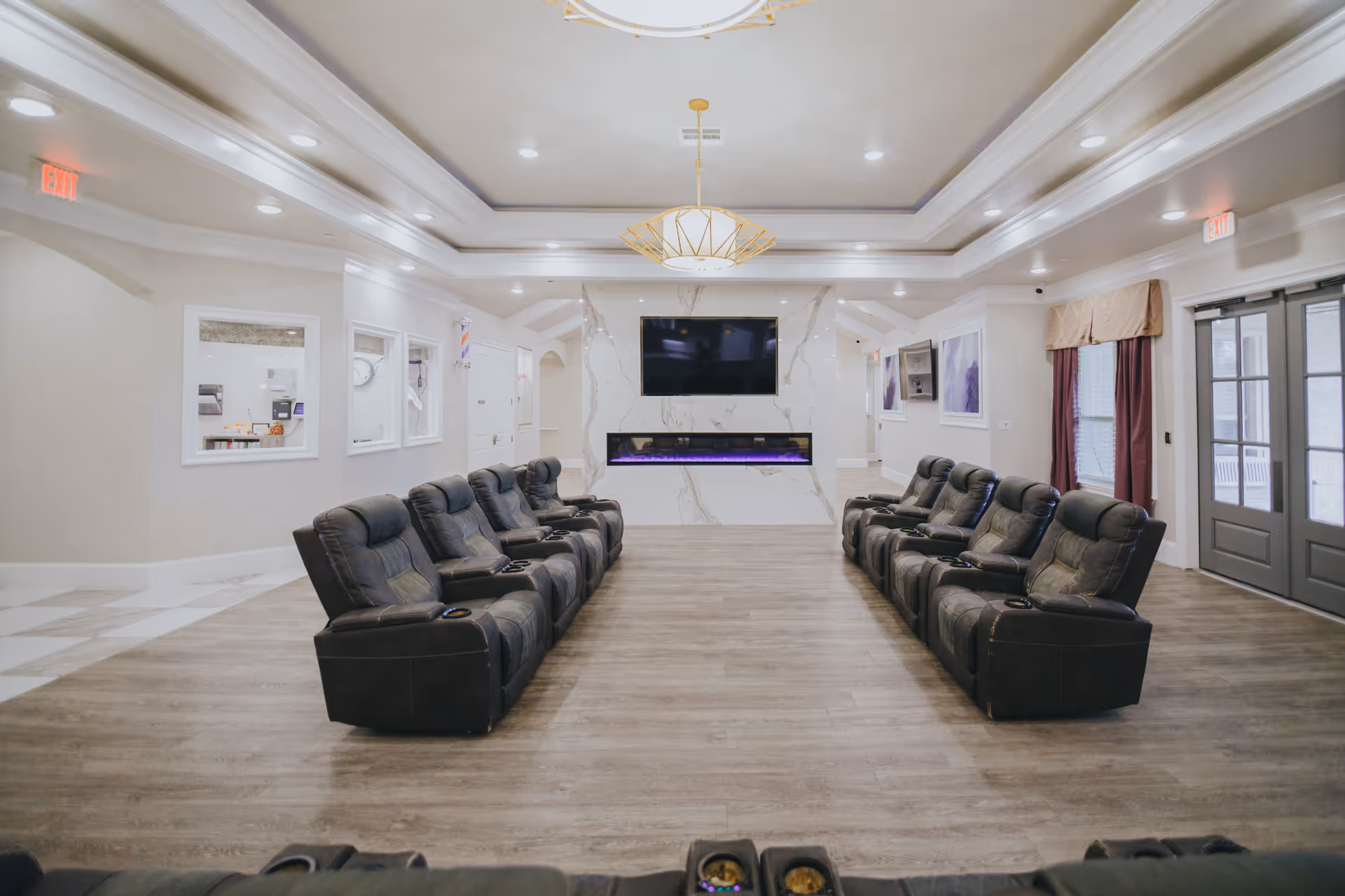 A spacious and well-lit common area with two rows of black reclining chairs facing a large flat-screen TV mounted above a modern electric fireplace with purple flames. The room has light-colored walls, a decorative ceiling light fixture, and large windows with curtains on the right side. There are exit signs and framed pictures on the walls.