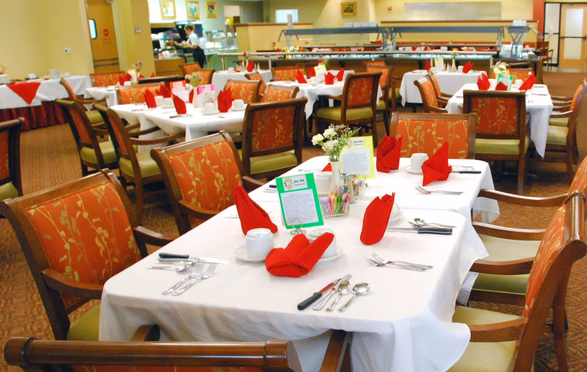 A dining room in a retirement community with multiple tables covered in white tablecloths, each set with red folded napkins, white cups, silverware, and small flower arrangements. The chairs have wooden frames with orange and green patterned upholstery. In the background, a kitchen or serving area is visible with a person working.
