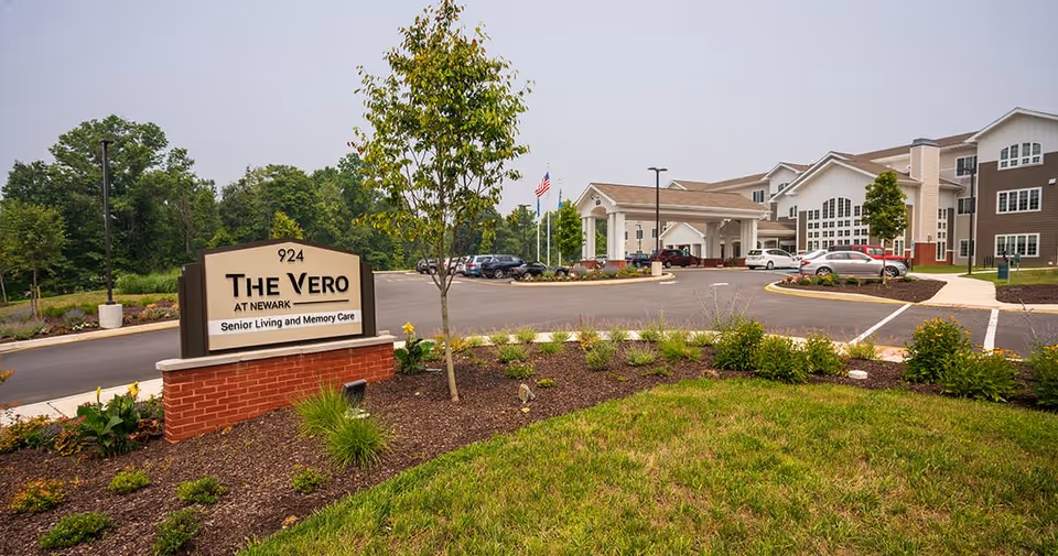 Exterior view of The Vero at Newark senior living and memory care facility showing the entrance driveway, parking lot with several cars, landscaped garden with small trees and shrubs, and a large sign with the facility name and address.