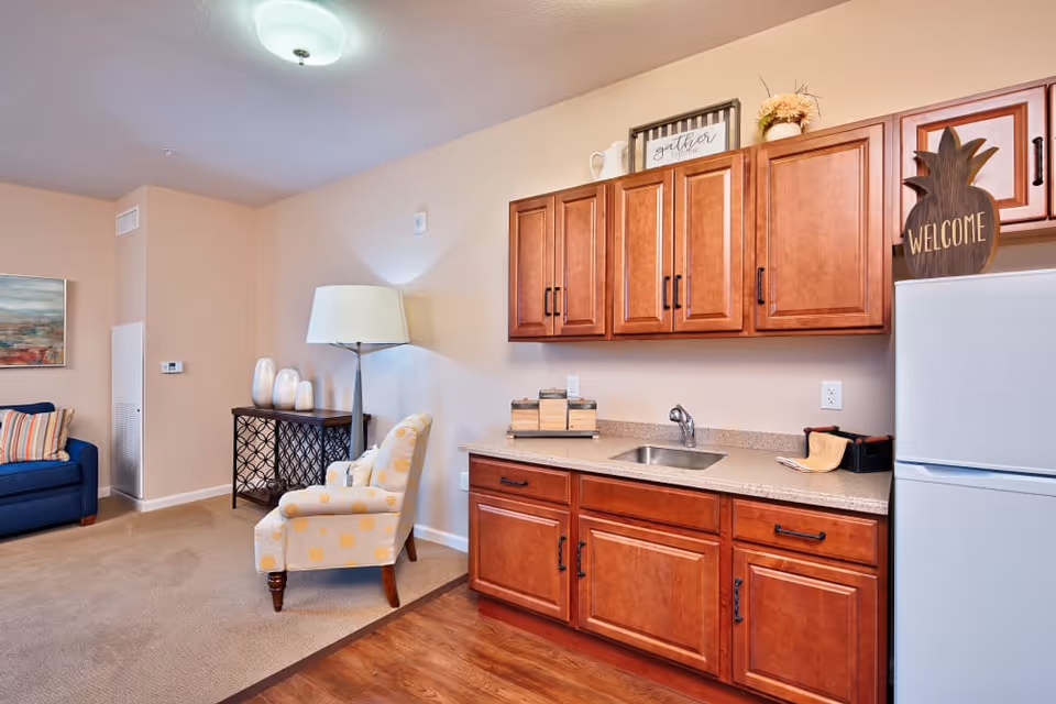 Studio-style living area with a kitchenette featuring wood cabinets, a sink and refrigerator, plus a patterned armchair, couch and floor lamp.