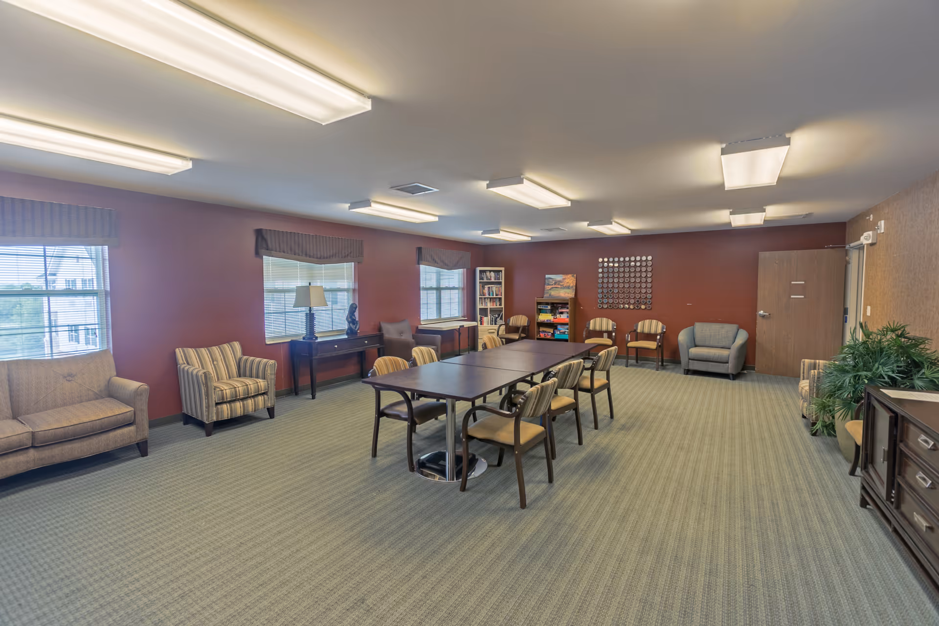 A spacious common room with a long table surrounded by chairs in the center. The room has multiple armchairs and sofas along the walls, windows with blinds, a bookshelf filled with games and books, and a decorative wall piece. The carpet is greenish, and the walls are painted a warm reddish color. The ceiling has several fluorescent lights.