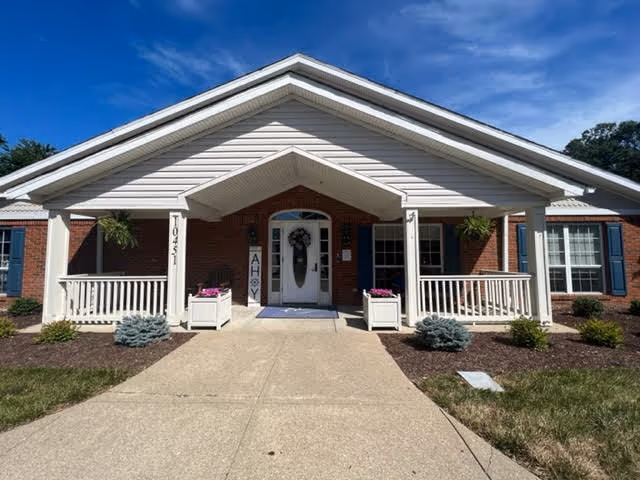 Front exterior view of a single-story brick building with white trim and a covered entrance. The entrance has a white door with a wreath, flanked by two white railings and planters with flowers. The building has blue shutters on the windows and a clear blue sky above.