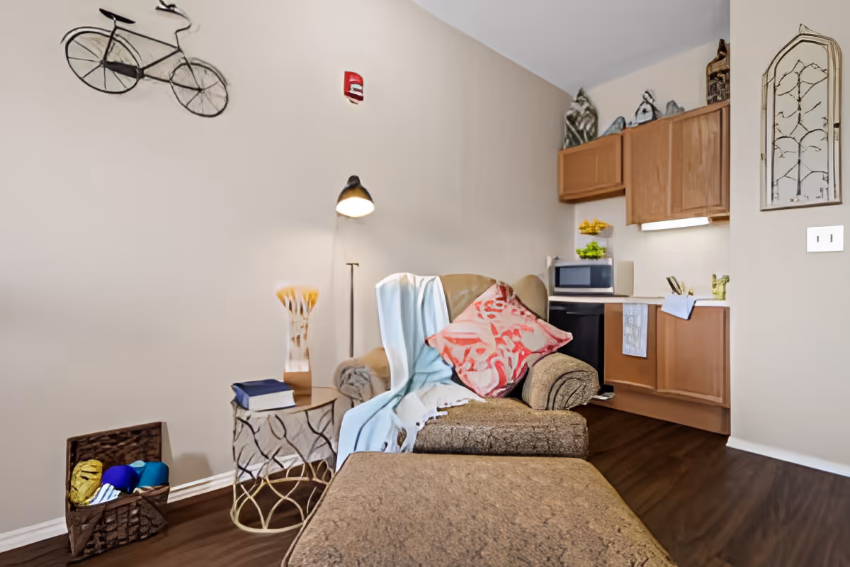 A cozy living area with a brown armchair and matching ottoman, adorned with a red patterned pillow and a light blue throw blanket. Next to the chair is a small round side table with a book and a decorative lamp. Behind the chair is a floor lamp and a wall-mounted metal bicycle decoration. In the background, there is a small kitchen area with wooden cabinets, a microwave, and a countertop with a towel hanging. The floor is dark wood, and the walls are painted beige.