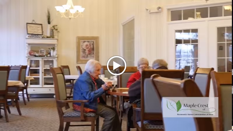 A dining room in a senior living facility with elderly residents sitting around a table, engaged in conversation. The room has multiple tables and chairs, a chandelier, a cabinet with decorative items, and framed artwork on the wall.